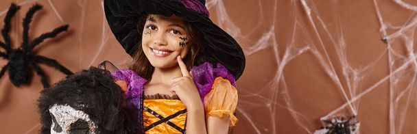 cerrar chica alegre en sombrero de bruja con juguete espeluznante tocando mejilla y sonriendo, Halloween, pancarta - Foto, imagen