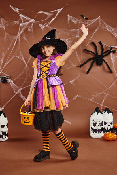 smiley preteen kid holds bucket of sweets and waving, brown backdrop with spiderweb, Halloween - Photo, Image