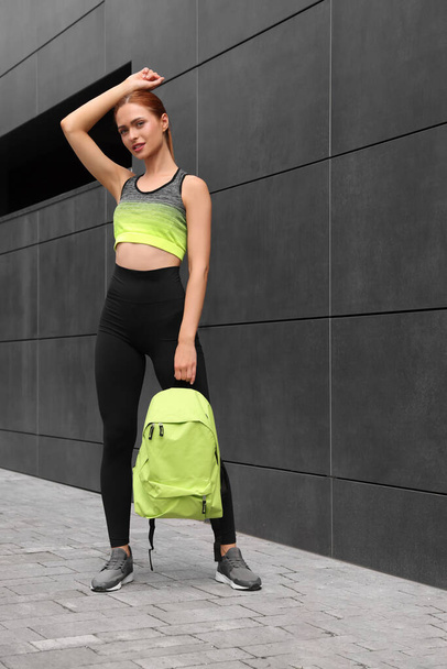 Beautiful woman in gym clothes posing with stylish backpack near dark grey wall on street - Фото, зображення