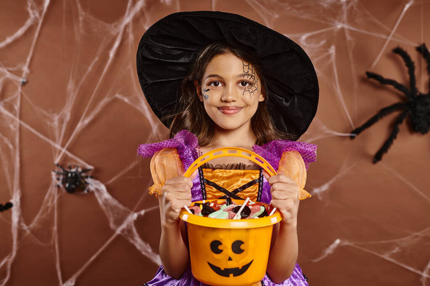 chica feliz en sombrero de bruja con maquillaje spiderweb celebración cubo de dulces en marrón con red de araña - Foto, imagen