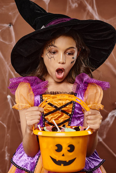 шокированный ребенок в шляпе ведьмы и костюме Хэллоуина смотрит на сладости в ведре на коричневом фоне - Фото, изображение