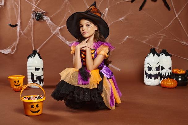 girl in witch hat and Halloween costume looking at camera near sweets in bucket and spooky decor - Photo, Image