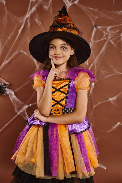 happy girl in witch hat and Halloween costume near cobwebs on brown backdrop, spooky season - Photo, Image