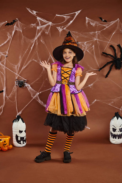 gelukkig meisje in heks hoed en Halloween kostuum gebaren in de buurt spinnenwebben op bruin, griezelig seizoen - Foto, afbeelding
