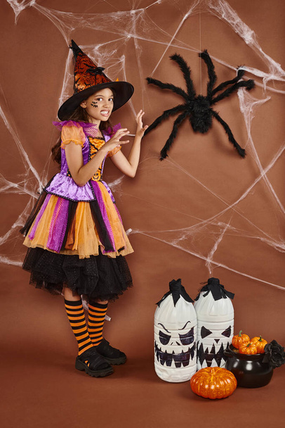 spooky girl in witch hat and Halloween costume growling near fake spider on brown background - Photo, Image