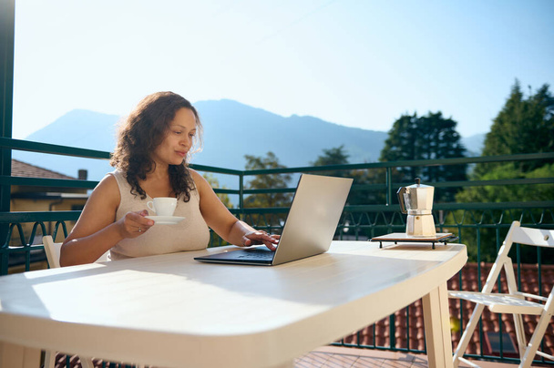 Szczęśliwa pewna siebie kobieta przedsiębiorca pije kawę i przy użyciu laptopa sprawdza pocztę, pracuje zdalnie, planuje projekty biznesowe, telekomunikacja z domu taras z widokiem na hipnotyzujący krajobraz - Zdjęcie, obraz