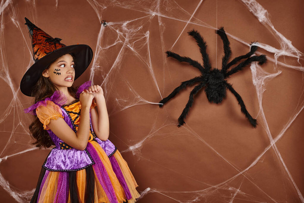 girl in witch hat and Halloween costume grimacing near fake spider and cobwebs on brown background - Photo, Image