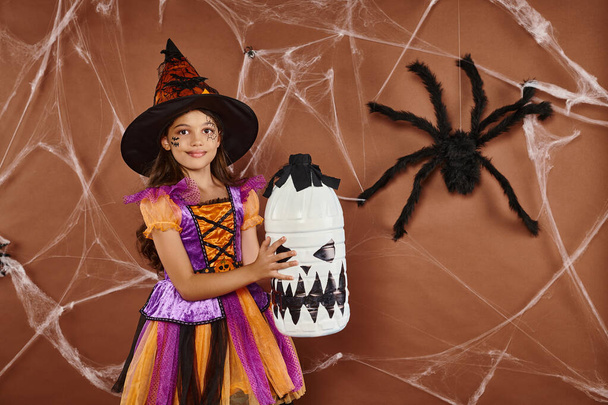 happy girl in witch hat and Halloween costume standing with spooky decor on brown background - Photo, Image