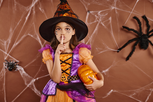 ragazza in cappello da strega e costume di Halloween in piedi con zucca e mostrando silenzio su sfondo marrone - Foto, immagini
