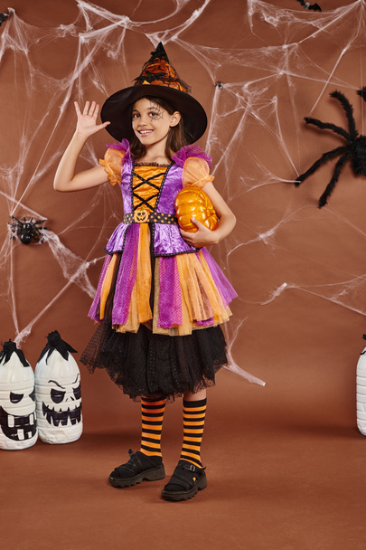 joyful girl in witch hat and Halloween costume standing with pumpkin and waving hand on brown - Photo, Image