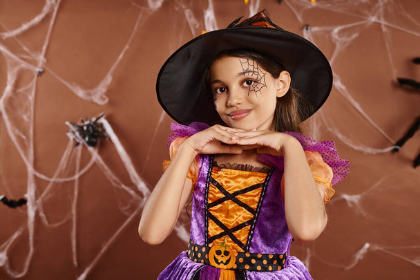 cute girl in Halloween witch costume and pointed hat smiling on brown backdrop, spooky season - Fotografie, Obrázek