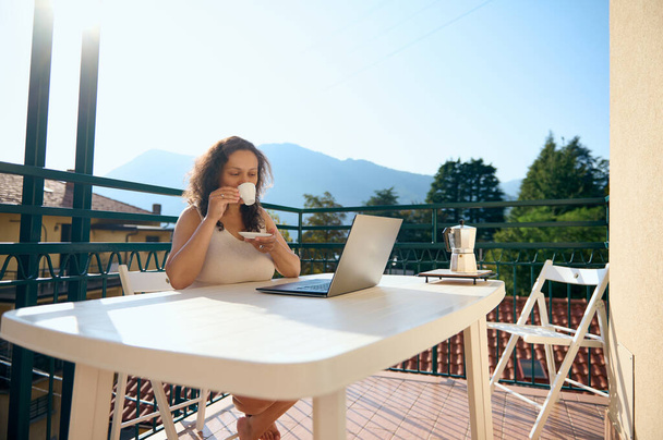 Authentisches Porträt einer entspannten, wunderschönen multiethnischen Geschäftsfrau, die Kaffee trinkt, während sie vom Balkon aus ferngesteuert am Laptop arbeitet und ein neues Geschäftsprojekt plant. Menschen. Unternehmen, Start-up - Foto, Bild