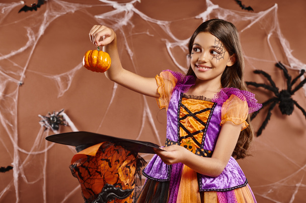 positive child in Halloween witch costume holding pumpkin near pointed hat on brown background - Photo, Image
