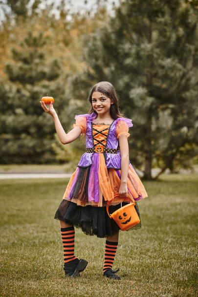 cheerful girl in Halloween costume holding pumpkin and bucket of candies on green grass - Photo, Image