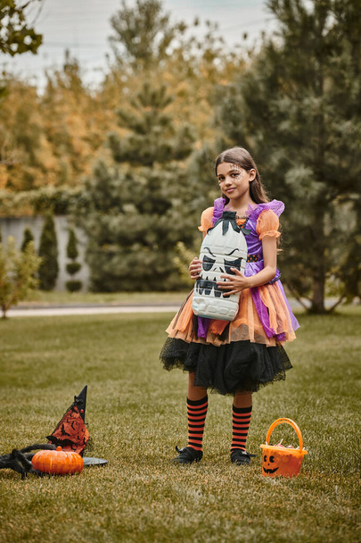 fille en costume d'Halloween tenant la décoration effrayante bricolage près de la citrouille, chapeau pointu et seau de bonbons - Photo, image