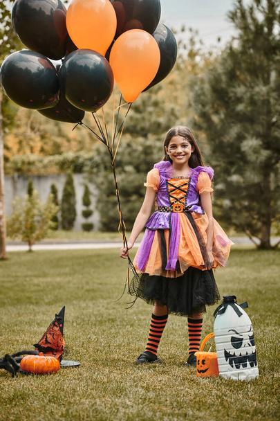 šťastná dívka v halloweenském kostýmu drží balónky v blízkosti dýně, špičatý klobouk a kbelík na sladkosti na trávě - Fotografie, Obrázek
