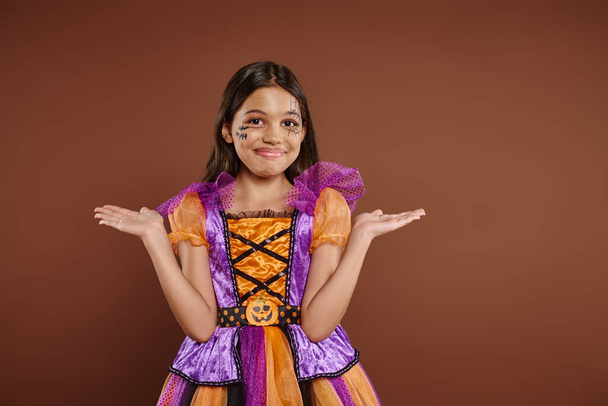 Verwirrtes Mädchen im Halloween-Kostüm mit Spinnennetz-Make-up lächelnd und gestikulierend vor braunem Hintergrund - Foto, Bild