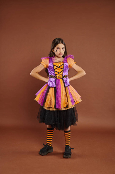 chica disgustado en disfraz de Halloween vestido de pie con las manos en las caderas en el fondo marrón, 31 de octubre - Foto, Imagen