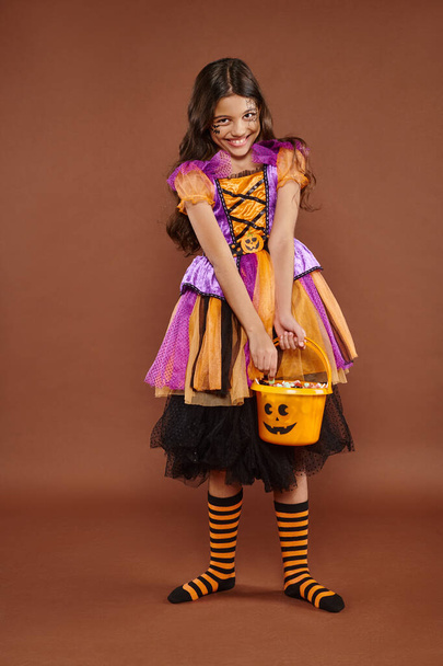 heerlijk meisje in Halloween kostuum holding emmer met snoepjes en houden rok op bruine achtergrond - Foto, afbeelding