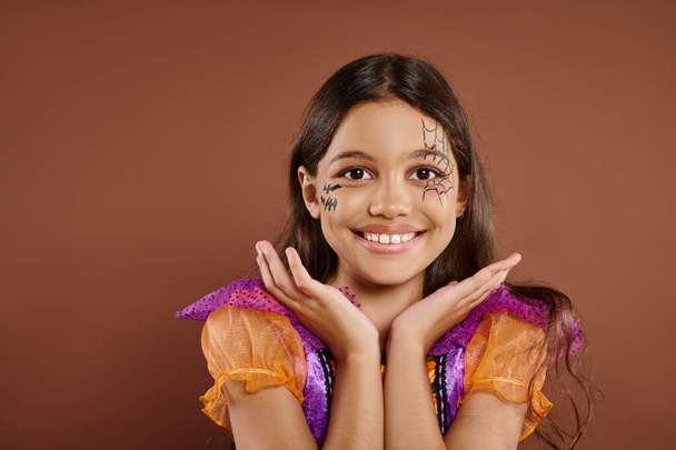 cheerful girl in Halloween costume and spiderweb makeup gesturing on brown backdrop, trick or treat - Photo, Image