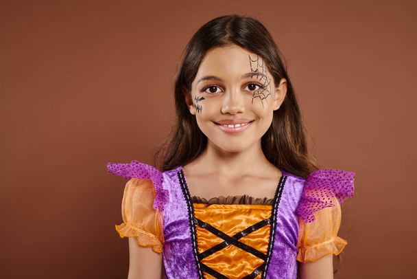 adorable girl in Halloween costume and spiderweb makeup smiling on brown backdrop, trick or treat - Photo, Image