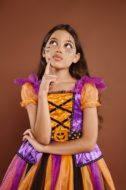 thoughtful girl in colorful costume with Halloween makeup looking away on brown background, October - Photo, Image