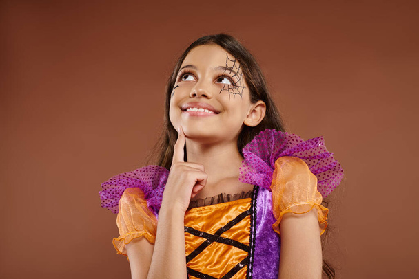 zasněný dívka v barevném kostýmu s Halloween make-up odvrací na hnědém pozadí, šťastný obličej - Fotografie, Obrázek