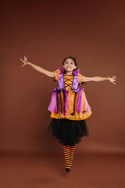 glückliches Mädchen im bunten Halloween-Kostüm, das in die Kamera blickt und auf braunem Hintergrund springt, schwebt - Foto, Bild
