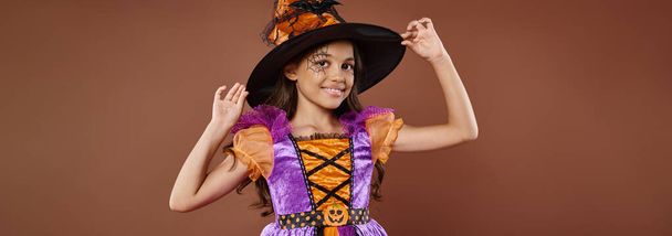 gelukkig meisje in Halloween kostuum en puntige hoed poseren op bruine achtergrond, kleine heks banner - Foto, afbeelding