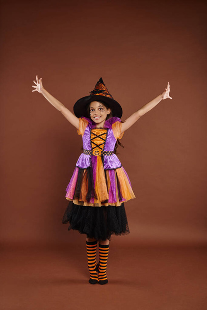 excited girl in Halloween costume and pointed hat standing with raised hands on brown background - Photo, Image
