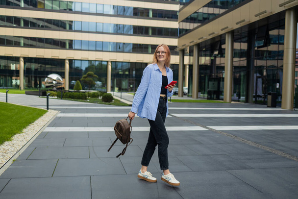 Χαρούμενη ευτυχισμένη κομψή γυναίκα χρησιμοποιώντας smartphone online app, ενώ στέκεται κοντά στο εμπορικό κέντρο. Ευτυχισμένη γυναίκα με μπλε πουκάμισο περπατά στο δρόμο κοιτάζοντας το smartphone της.Μια συναρπαστική εφαρμογή που χρησιμοποιείται για - Φωτογραφία, εικόνα