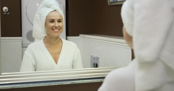 Завернутая в халат, белая женщина смотрит в зеркало с довольной улыбкой. Ее отражение излучает позитив, захватывая момент самооценки и уверенности. - Кадры, видео