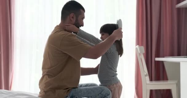 Ein Vater verteilt Multitasking, während er seiner Tochter liebevoll die Haare kämmt, seine Berührung sanft und beruhigend. In einer herzerwärmenden Geste zieht er sie dann in eine warme Umarmung. - Filmmaterial, Video