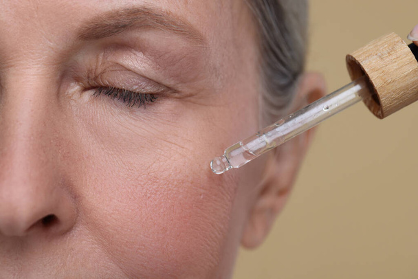 Senior woman applying cosmetic product on her aging skin against beige background, closeup. Rejuvenation treatment - Φωτογραφία, εικόνα