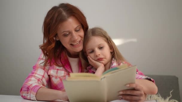 Positiivinen valkoihoinen nainen, joka lukee ääneen tytön kuuntelutarinaa. Hymyilevä kaunis äiti nauttii harrastus söpö viehättävä tytär kotona istuu pöydässä - Materiaali, video