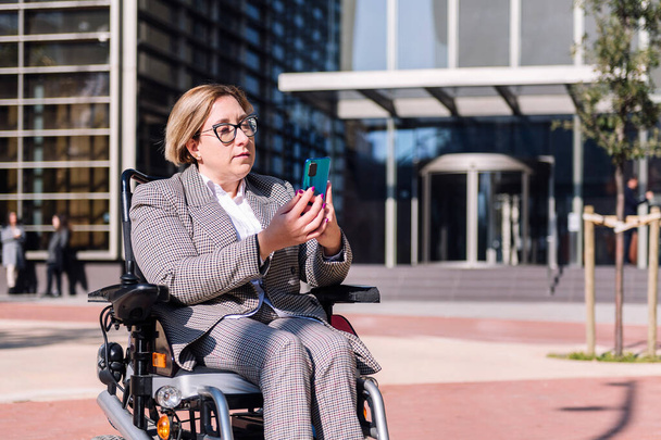 businesswoman using wheelchair using mobile phone in front of an office building in the financial district, concept of diversity and technology of communication - Photo, image
