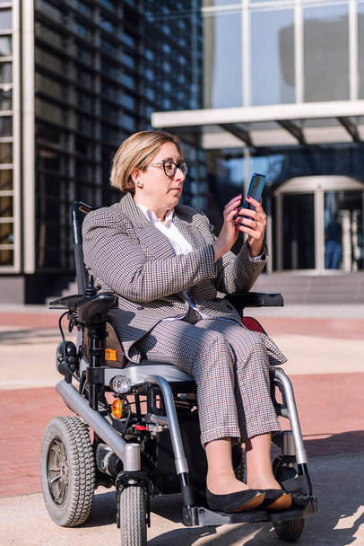 businesswoman using wheelchair typing a message on mobile phone in front of an office building in the financial district, concept of diversity and technology of communication - Valokuva, kuva