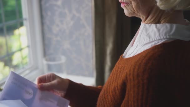 Senior woman standing by window at home opening UK energy bill during cost of living crisis looking worried - shot in slow motion - Filmmaterial, Video