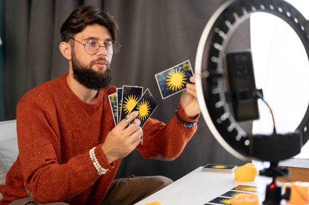 Online astrology. Male fortune teller is use a smartphone and ring light, reads tarot cards for predictions, telling customers by providing services via mobile phones. Concept of divination. Copy space - Photo, Image
