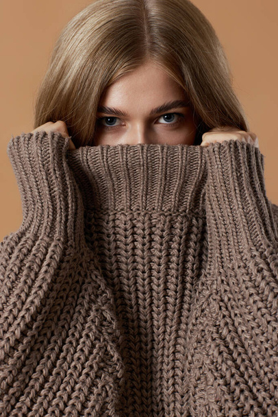 beautiful blonde woman covering face with turtleneck knitted sweater on beige background. autumn style - Фото, изображение