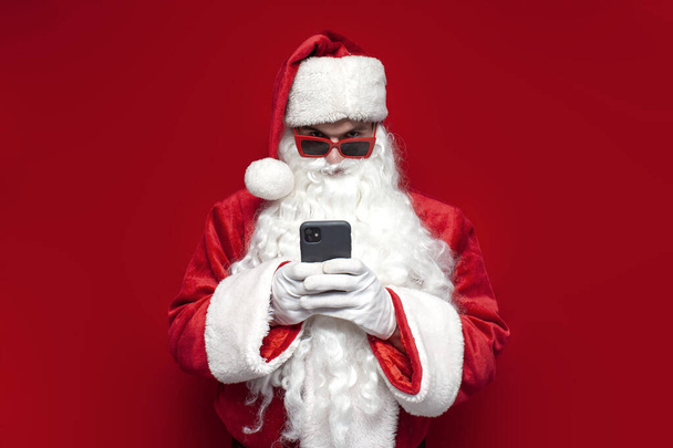 Санта-Клаус в шляпе и праздничных очках использует смартфон на красном фоне, мужчина в костюмах Санта-Клауса типа онлайн по телефону на Рождество - Фото, изображение
