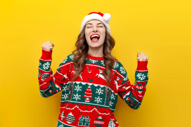 jeune fille en pull de Noël et santa claus chapeau célèbre la victoire et le succès sur fond jaune isolé, femme gagnante se réjouit de la chance dans les vêtements de la nouvelle année - Photo, image
