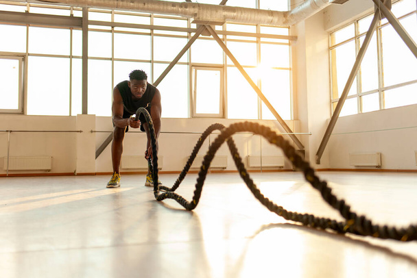 Spor giyinen Afrikalı Amerikalı bir adam spor salonunda sabah antrenman yapıyor, adam halatları sallıyor ve spor salonunda spor yapıyor. - Fotoğraf, Görsel