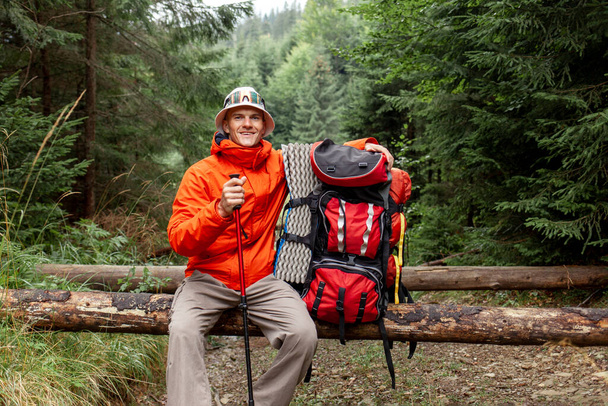 чоловік турист з рюкзаком і пішохідним спорядженням сидить відпочиває в лісі, хлопець в помаранчевій куртці на гірському поході робить перерву - Фото, зображення