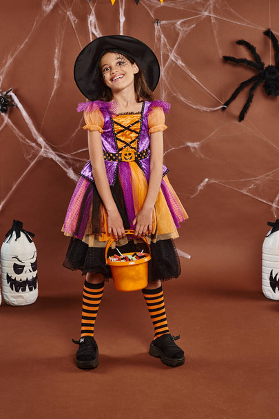 happy girl in witch hat and Halloween costume holding bucket with sweets near diy spooky decor - Photo, Image
