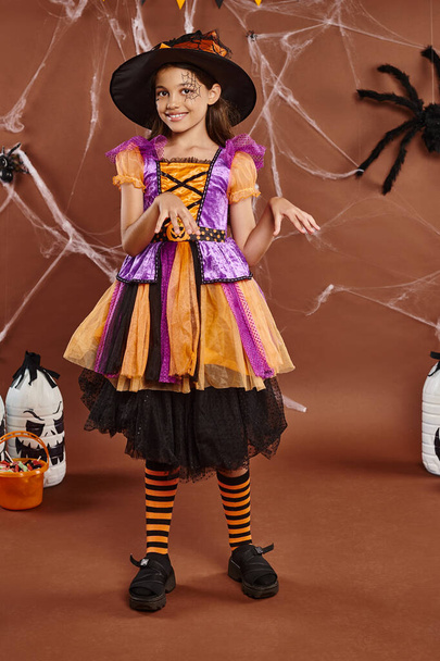 fille gaie en chapeau de sorcière et robe dansant près du seau avec des bonbons sur brun, concept d'Halloween - Photo, image