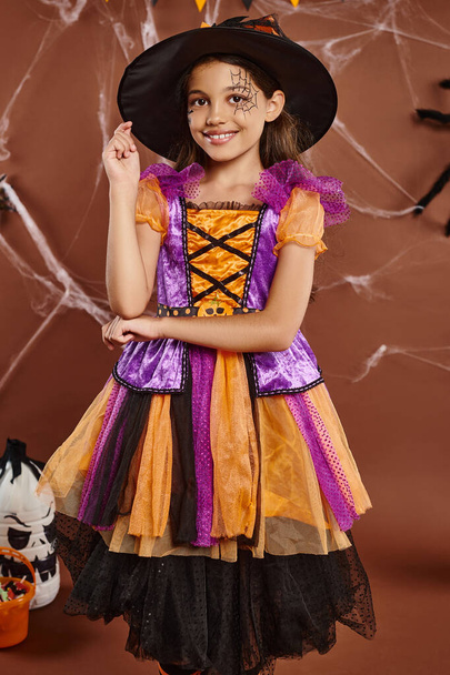 chica positiva en traje de bruja y sombrero puntiagudo sonriendo sobre fondo marrón, concepto de Halloween - Foto, Imagen