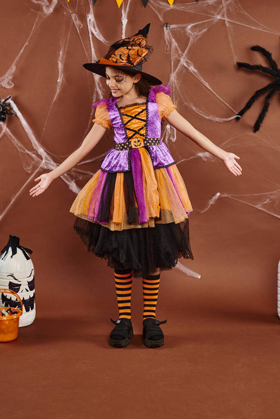 pozitivní dívka v kostýmu čarodějnice a špičatý klobouk gestikulující poblíž kbelíku na sladkosti, Halloween koncept - Fotografie, Obrázek
