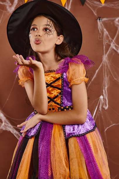 linda chica en traje de bruja y sombrero puntiagudo enviando beso de aire sobre fondo marrón, concepto de Halloween - Foto, Imagen