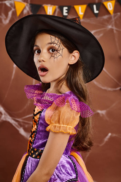 sorprendido pequeña bruja en disfraz de Halloween y sombrero puntiagudo sobre fondo marrón, chica sorprendida - Foto, Imagen